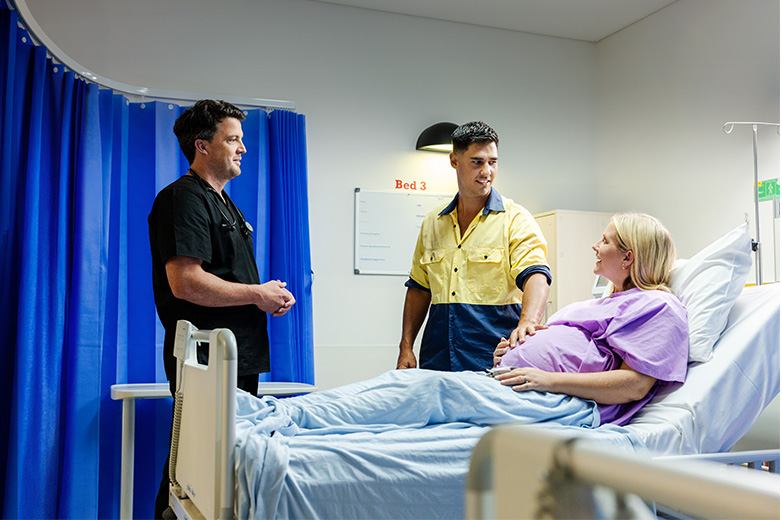 Pregnant woman in a hospital bed speaking with a doctor and a man in a high-visibility shirt beside the bed.