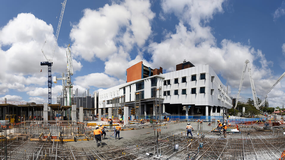 Wide view of the Ipswich Hospital expansion site with cranes, scaffolding, and workers preparing steel framework for new building foundations.