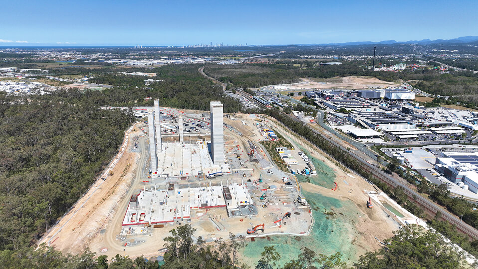 Wide aerial view of the Coomera Hospital site with concrete structures rising and surrounding landscape and suburbs visible.
